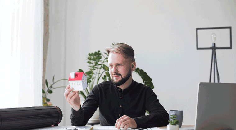 Um homem sentado em uma mesa segurando uma casa em miniatura